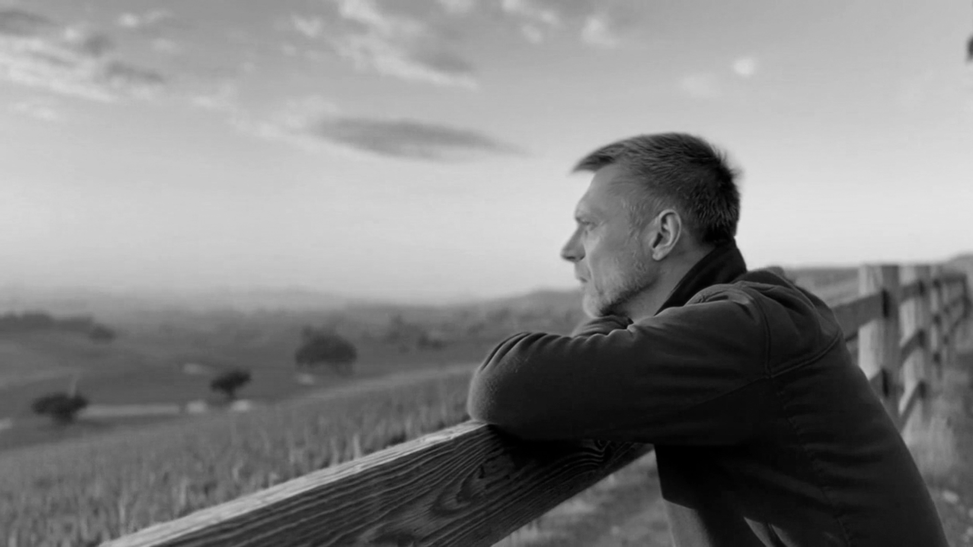 A black and white portrait of a man leaning on a wooden fence, looking out over an open landscape in quiet contemplation.