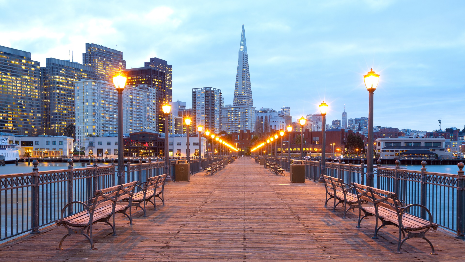 A quiet city pier at dusk with empty benches and warm lights leading toward the skyline, suggesting patience and forward movement.