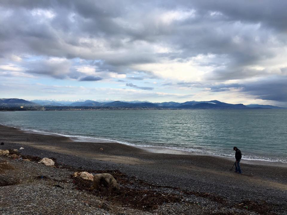 A rocky beach in Antibes under a cloudy sky, with waves rolling in and a lone figure walking along the shoreline