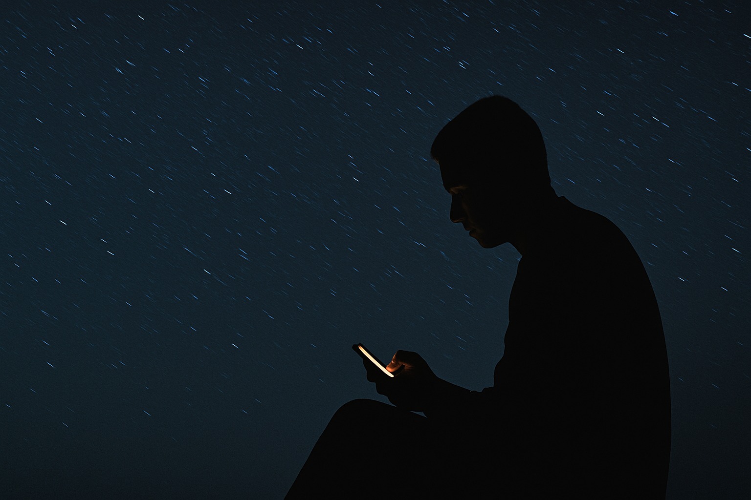 A solitary figure illuminated by a phone under a star-filled night sky, suggesting quiet contemplation within vast motion